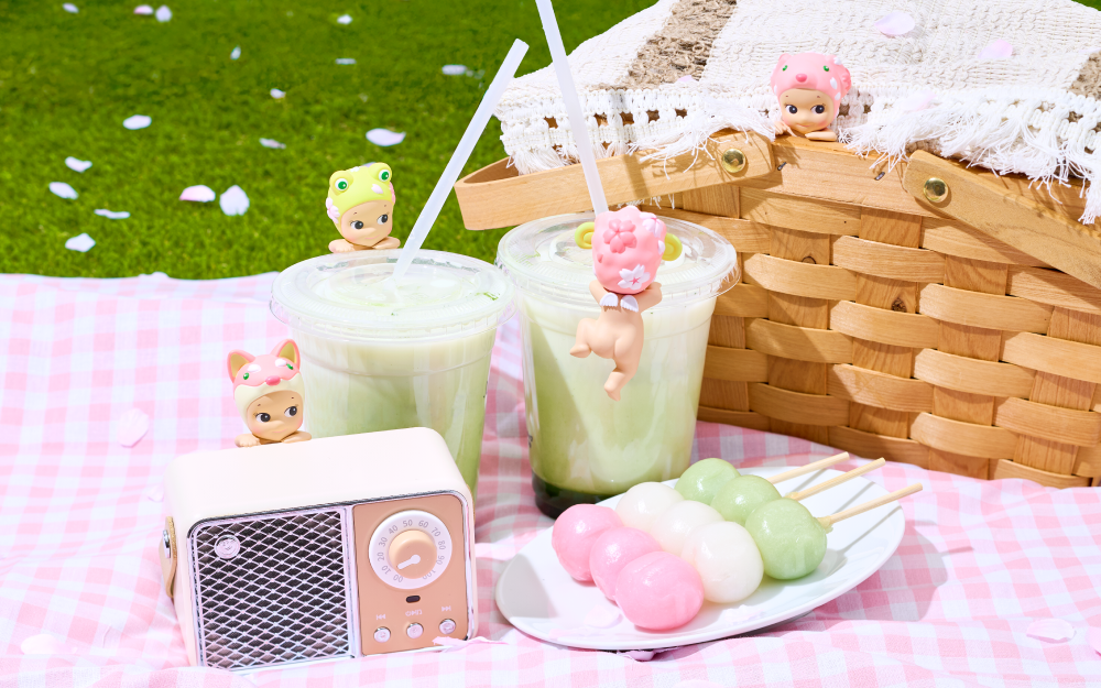Cherry Blossom HIPPERS product image shows a picnic on the grass with the Dalmatian HIPPER peeking out from the picnic basket. On the picnic blanket, the Shiba HIPPERs perches on a radio, beside the Frog HIPPER facing out on an iced matcha, beside the Sheep HIPPER facing away on an iced matcha, placed behind a plate of hanami dango.