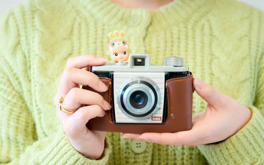 Person in a light green sweater is holding a vintage-style camera with the Peacock Cherry Blossom HIPPER peeking out over the top