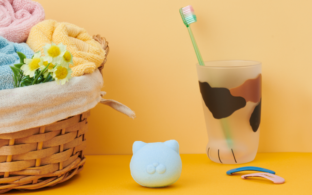 Product image for Nekomote Bath Bomb- Rosemary scent. Set against a light yellow background, atop a bright yellow counter, center image shows the blue bath bomb product. To the left of the bath bomb sits a basket of towels, to the right a calico patterned cup with toothbrush, and hairclips