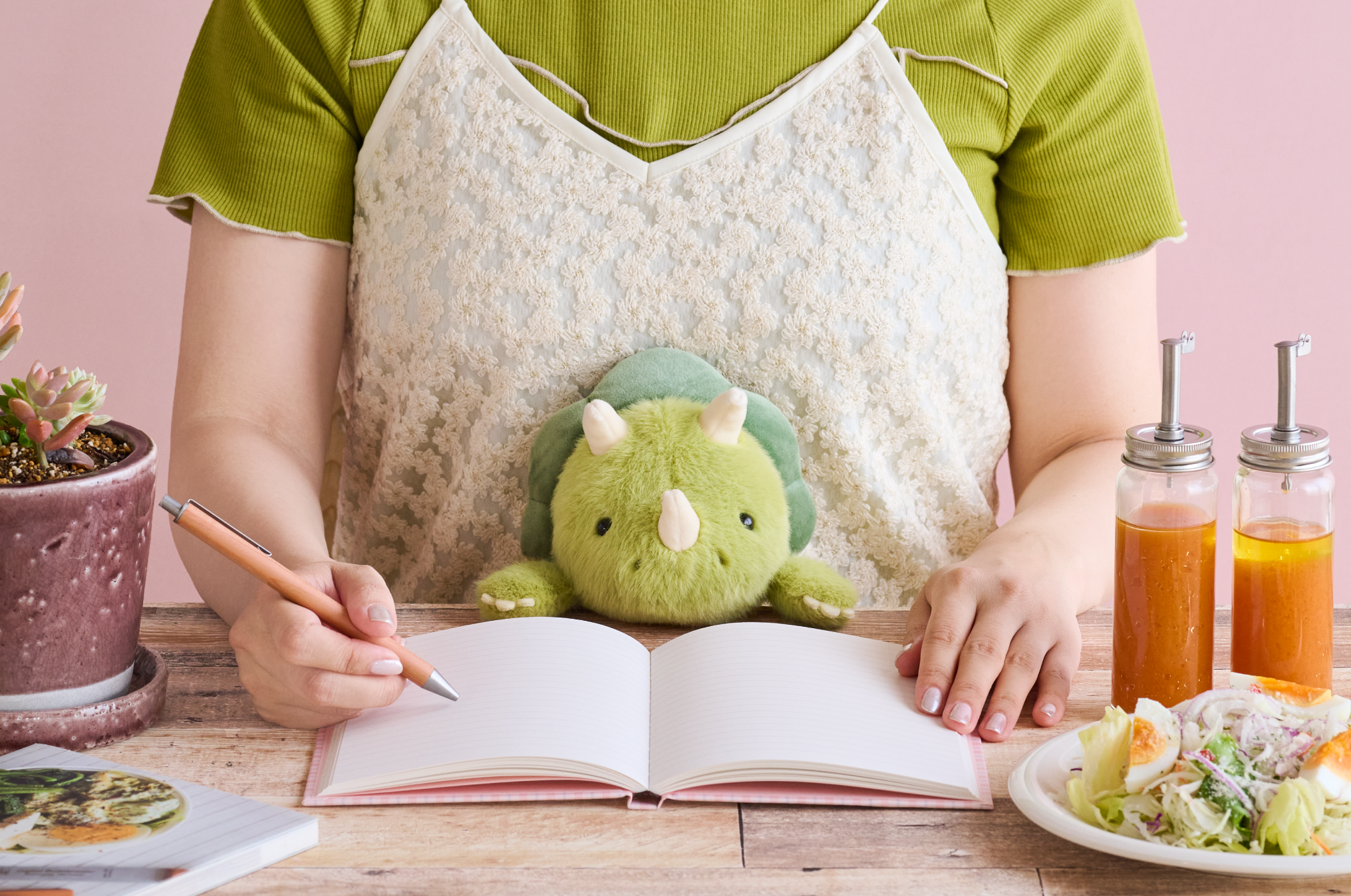 Image set on pink background. Figure in layered green and white shirts sits at a desk. Light and dark green Triceratops Posture Pal supports figure as they write in an open notebook. A potted succulent and book are on one side of figure, with a salad and two salad dressings set on the other.